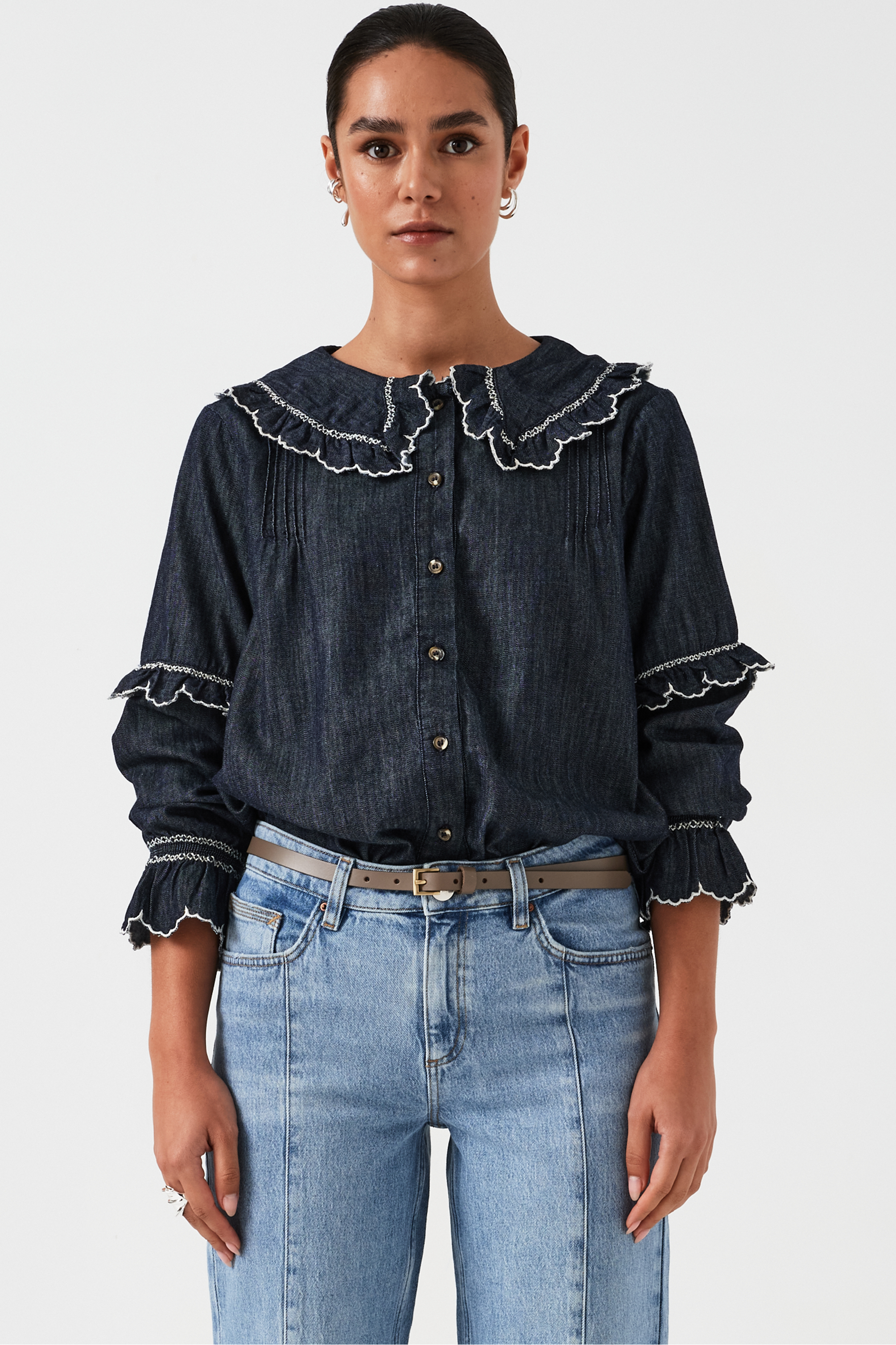 A woman wearing a navy denim blouse with a white trim frill collar and frill sleeves, paired with blue jeans on a white background. The blouse is lightweight and made of organic cotton.