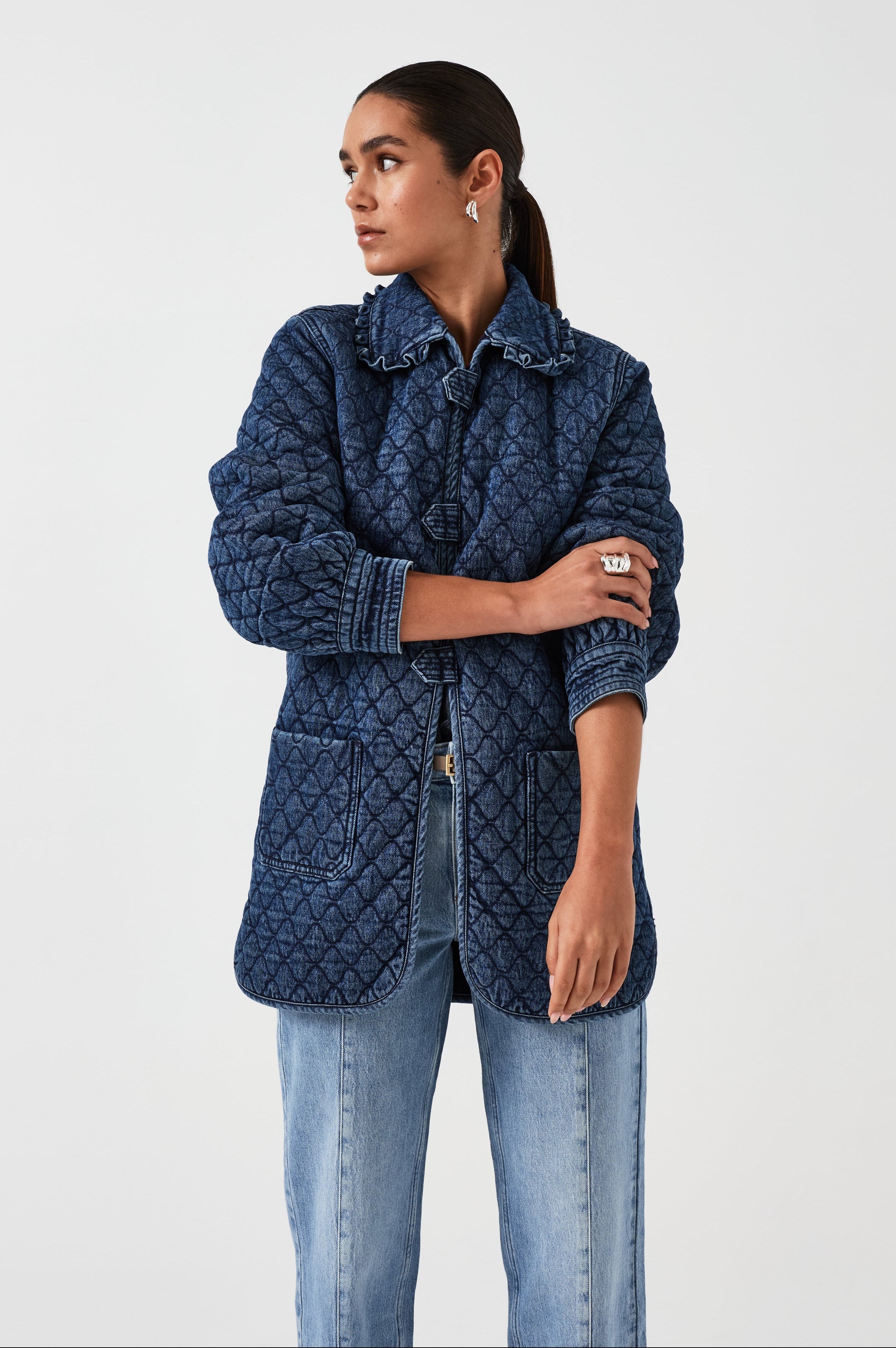woman wearing a blue quilted jacket with a frill collar, tab closures, and two large pockets on a white background