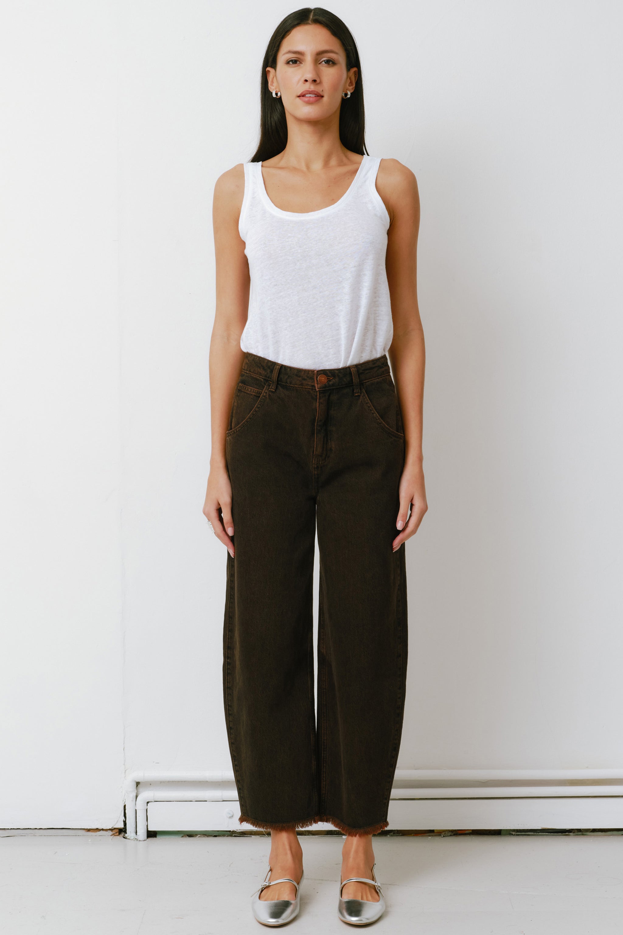 woman standing against a white background wearing high-rise dark brown cacao-colored carpenter barrel leg jeans with a frayed hem and a white top.