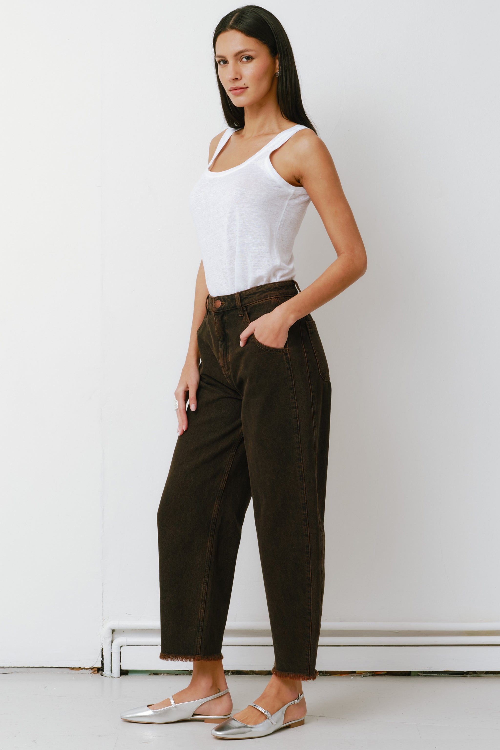 woman standing against a white background wearing high-rise dark brown cacao-colored carpenter barrel leg jeans with a frayed hem and a white top.
