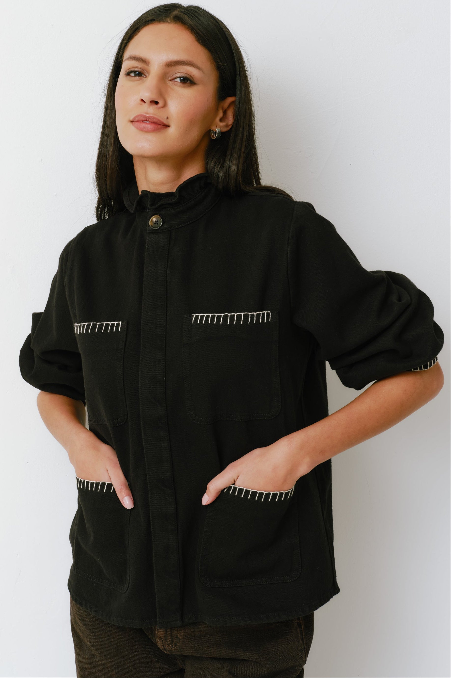 A woman wearing a black denim jacket with a frill collar and our signature white blanket stitching on four large patch pockets, paired with brown jeans against a white backdrop