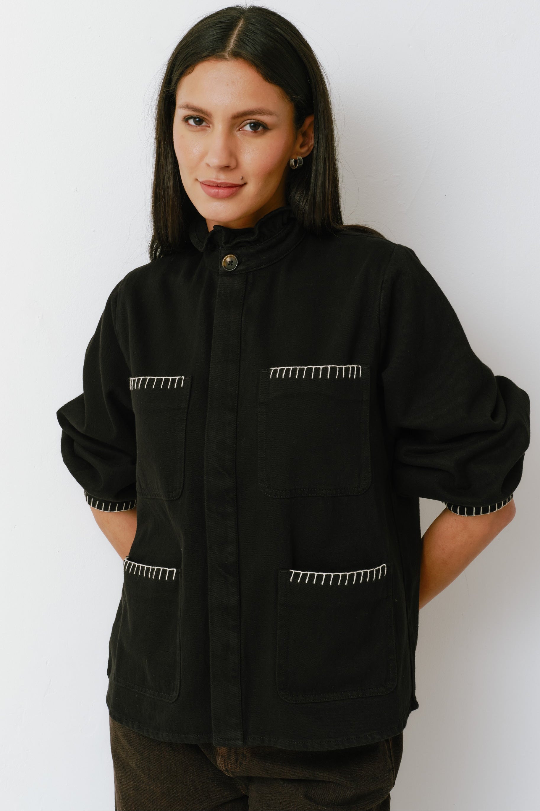 A woman wearing a black denim jacket with a frill collar and our signature white blanket stitching on four large patch pockets, paired with brown jeans against a white backdrop