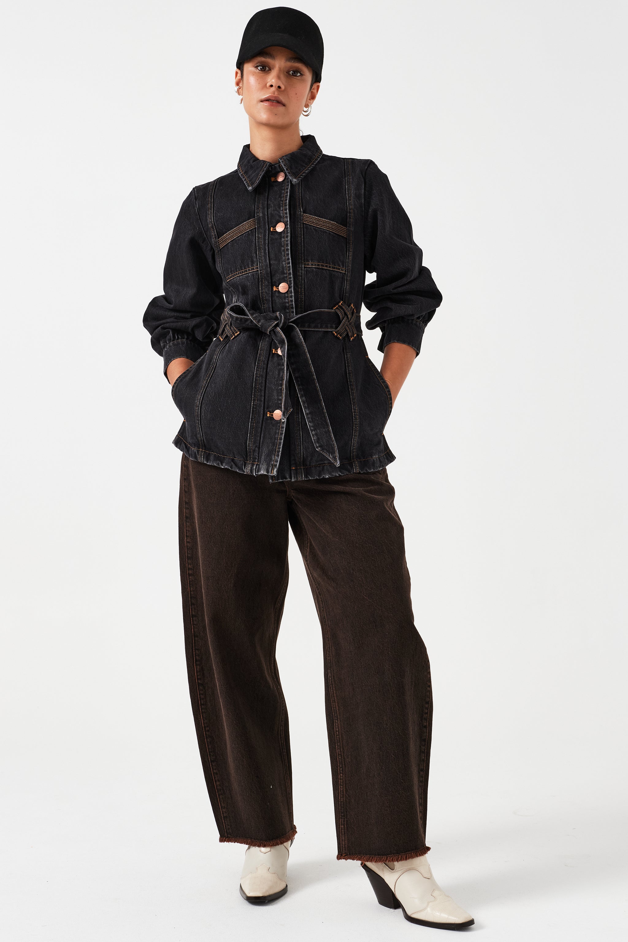 woman standing against a white background wearing high-rise dark brown cacao-colored carpenter barrel leg jeans with a frayed hem and dark wash denim jacket with a tie waist and a black hat