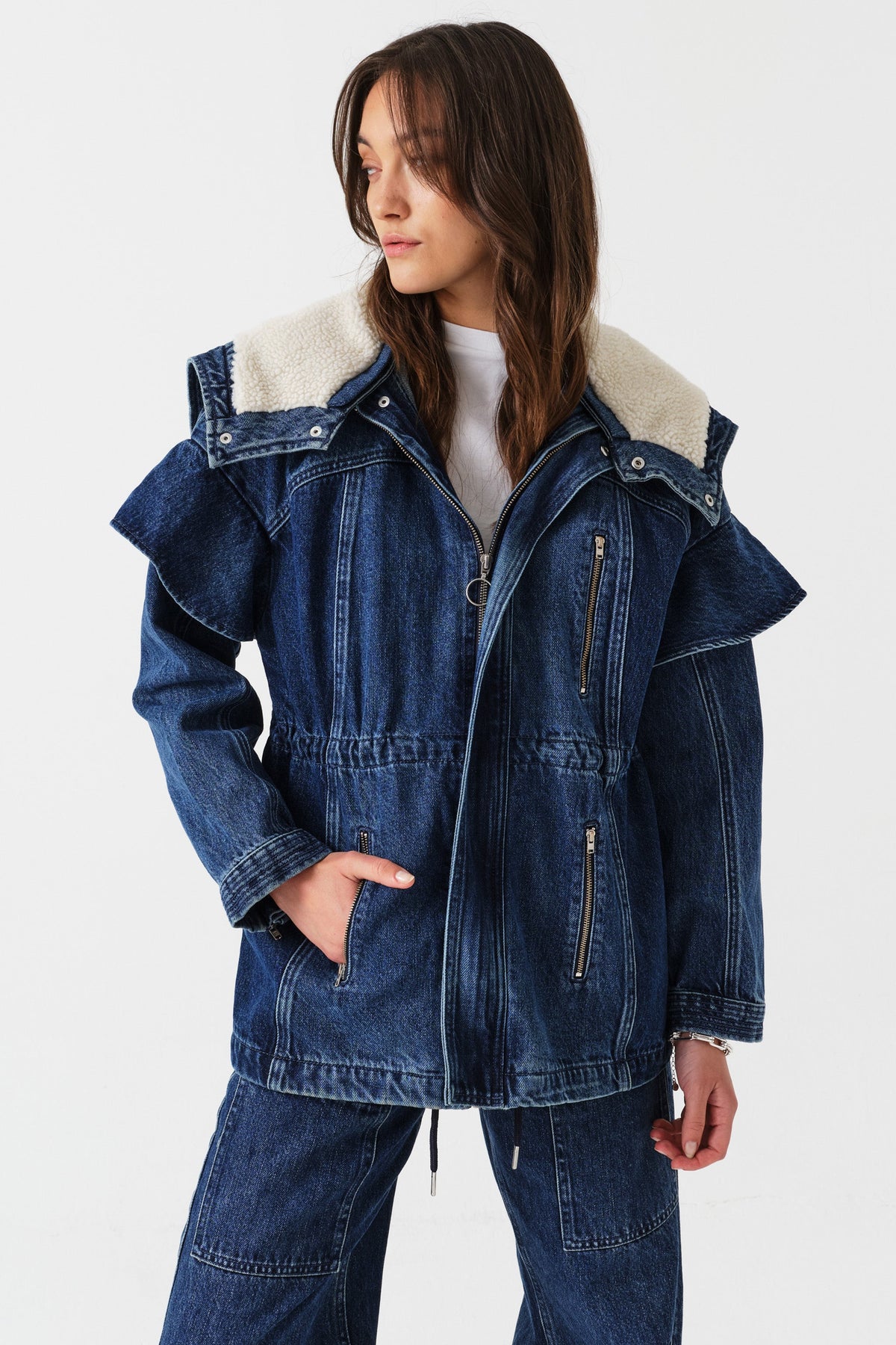 Close up of woman wearing a white shirt, dark blue denim jacket with a white sherpa lined removable hood, waist cinch, and zip pocket details with dark blue jeans with cargo zip pockets on a white background