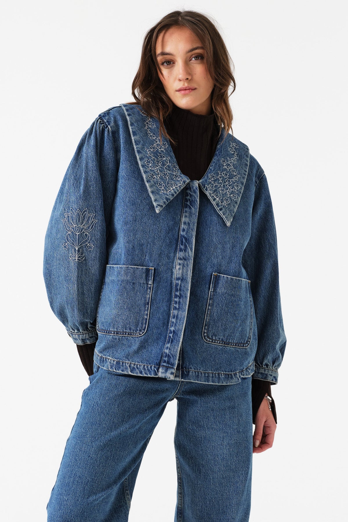 Woman wearing a blue denim jacket with balloon sleeves, embroidery, and pockets with blue jeans on a white background