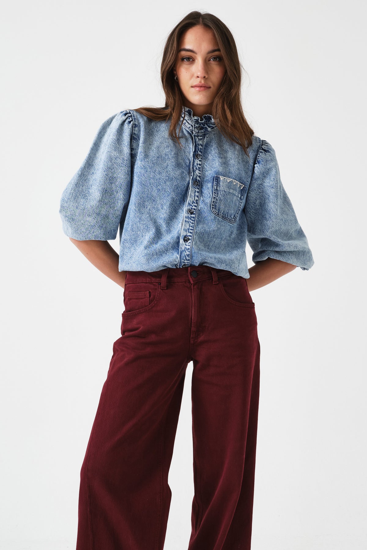 Woman wearing a denim shirt/ blouse with balloon sleeves and burgundy pants on a white background