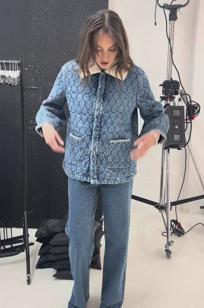 Woman wearing a blue quilted denim jacket with a removable white sherpa collar, large front pockets, and white embroidery details, and blue jeans